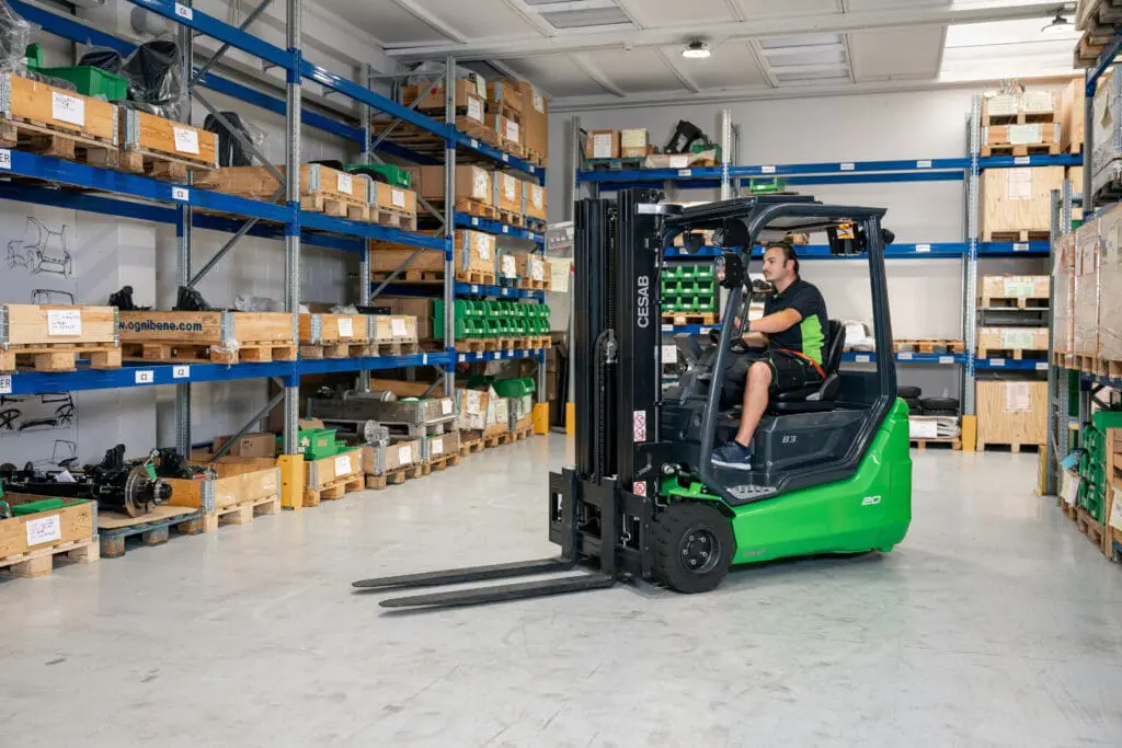 Driver on a green CESAB B3-20 forklift driving through a warehouse with blue shelving units. - Prince Maasdijk