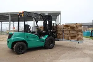 Green Prince Bulldog 25-E electric forklift with a stack of wooden pallets in an outdoor yard, left side view. - Prince Maasdijk